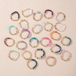 Puede incluir: Una colección de anillos de cuentas en varios colores, incluyendo negro, blanco, azul, morado y rosa. Cada anillo presenta una banda dorada texturizada. Los anillos están dispuestos sobre una superficie clara.