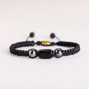 May include: A black macrame cord bracelet featuring a central rectangular black bead flanked by two metallic grey spherical beads. The adjustable band includes a small gold-toned accent bead and two smaller black beads at the cord ends.