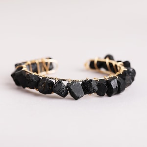 May include: A cuff bracelet featuring a series of raw, black, angular stone pieces secured to a thin, gold-toned metal wire frame. The stones are arranged in a continuous row along the curved structure, showcasing their natural, unpolished texture.
