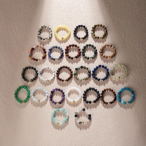 Puede incluir: Una colección de pulseras de cuentas en varios colores, incluyendo verde, azul, blanco y negro. Cada pulsera presenta una cuenta de acento dorada. Las pulseras están dispuestas sobre una superficie rosa claro.