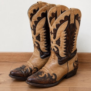 Könnte beinhalten: Ein Paar wadenhohe Cowboystiefel im zweifarbigen Design aus hellbraunem und dunkelbraunem Leder. Die Stiefel verfügen über aufwendige Ziernähte, spitz zulaufende Zehenpartien, Blockabsätze und Zuglaschen am oberen Schaftrand zum einfachen Anziehen.
