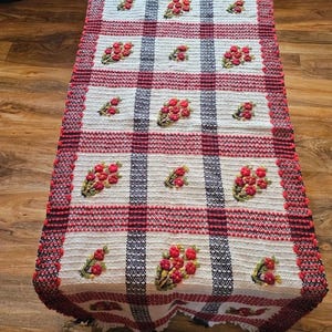 May include: A rectangular table runner featuring a white woven base with a grid pattern of dark blue and red stripes. The runner is decorated with embroidered clusters of red flowers and green leaves, finished with a red and dark blue textured border.