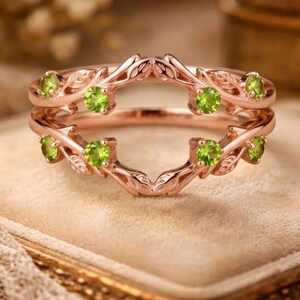 Può includere: Un anello in oro rosa con un design ispirato alla natura. L'anello presenta una doppia fascia con dettagli di foglie e viti ed è adornato da diverse pietre preziose rotonde verde chiaro. L'anello è posto su uno sfondo neutro.