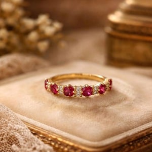 Può includere: Un anello d'oro con una serie di vivaci pietre preziose rosse e piccole pietre chiare. L'anello è presentato su uno sfondo morbido e neutro, evidenziando il suo design elegante. Le pietre sono uniformemente distanziate.