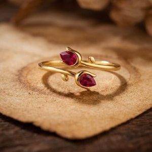 May include: A gold-colored ring featuring a floral design with two red gemstones. The ring's band curves around the finger, with each end forming a tulip-like shape holding a teardrop-shaped red stone. The ring is set against a neutral background.