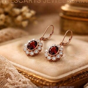 May include: A pair of rose gold drop earrings featuring oval-cut deep red garnet center stones surrounded by a halo of small, clear, round-cut white stones. The earrings are equipped with secure lever-back closures for comfortable wear.
