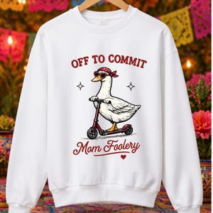 May include: White sweatshirt featuring a goose wearing sunglasses and a red bandana, riding a red scooter. The text reads "Off to Commit Mom Foolery" in maroon.