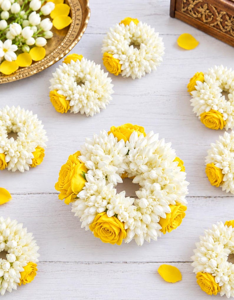 May include: A collection of circular floral garlands crafted from small white jasmine buds and accented with vibrant yellow roses. Each wreath features a dense, textured arrangement of white petals contrasted by bright yellow rose blossoms.
