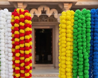 May include: Colorful garlands of flowers in white, red, yellow, green, and blue hang in front of a building with ornate architecture. The garlands are made of small, round, fluffy flowers, creating a vibrant display.