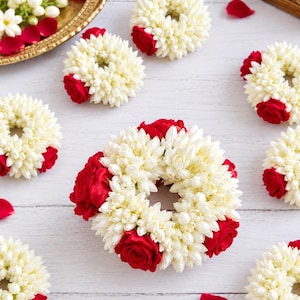 May include: A collection of circular floral garlands crafted from white jasmine buds and vibrant red rose blossoms. Each wreath features a dense arrangement of small white petals accented by deep red roses, creating a repeating pattern of floral rings.