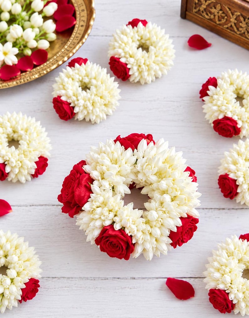 May include: A collection of circular floral garlands crafted from white jasmine buds and vibrant red rose blossoms. Each wreath features a dense arrangement of small white petals accented by deep red roses, creating a repeating pattern of floral rings.