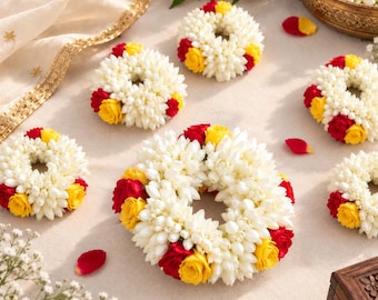 May include: A collection of circular floral garlands crafted from white jasmine buds, accented with vibrant red and yellow rose blossoms. These decorative wreaths are arranged in a cluster, featuring dense, layered petals and a fragrant, natural aesthetic.