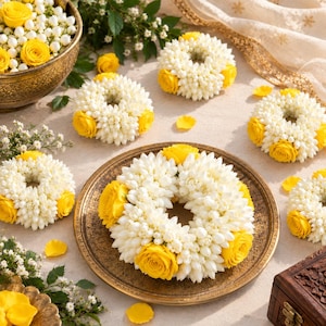 May include: A collection of circular floral garlands crafted from small white jasmine buds and bright yellow roses. Several garlands are arranged on a textured surface, with one large garland centered on a decorative bronze-toned metal plate.