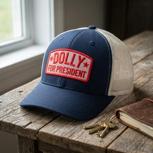 May include: A navy blue trucker cap with a white mesh back and a rectangular red patch on the front. The patch features the white text "DOLLY FOR PRESIDENT" with a small red star on each side of the word "DOLLY" and a red border.