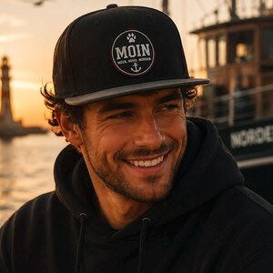 Könnte beinhalten: Schwarze Baseballkappe mit grauem Schirm. Die Kappe hat einen runden Aufnäher mit dem Wort "MOIN", einem Pfotenabdruck und einem Anker. Der Text "MEER. HUND. NORDEN." steht darunter.