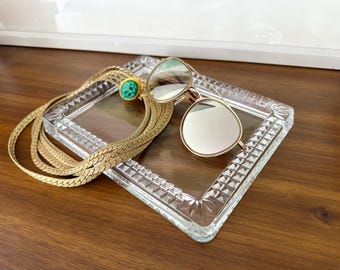 May include: A gold-toned necklace with a green gemstone pendant rests on a clear, square glass tray. Gold-rimmed sunglasses with reflective lenses are also on the tray. The tray sits on a dark wooden surface.