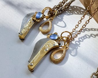 May include: Two gold-toned pocket knife pendants with silver blades and blue gemstone accents. Each pendant hangs from a gold chain, with a textured teardrop charm. The knives have ornate floral detailing.