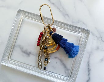 May include: A gold-toned metal oval carabiner keychain featuring a layered charm cluster. The assembly includes a multi-tiered blue thread tassel, a strand of red beads, a gold-toned chain, a small evil eye bead, and a textured gold-toned circular pendant.