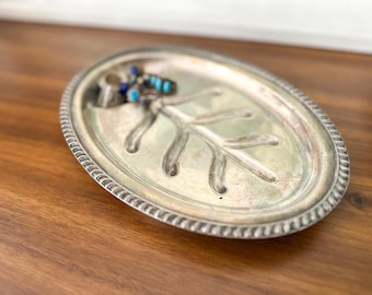 May include: Oval silver-toned jewelry tray with a decorative leaf design. The tray has a beaded edge and holds rings and turquoise and blue gemstone jewelry. The tray sits on a wooden surface.