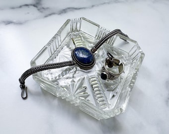 May include: A clear, faceted glass tray holding a silver bracelet with a dark blue oval stone, and two rings. One ring has a dark blue stone, and the other has a cluster of small stones. The tray sits on a white marble surface.