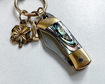 May include: A small pocket knife with a mother-of-pearl handle and gold accents attached to a keychain with a gold shamrock charm. The knife blade is silver, and the handle has iridescent colors.