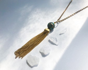 May include: A gold chain necklace with a turquoise and gold tassel pendant. The pendant is a round ball with intricate designs. The necklace is laying on a white surface with small white crystals.