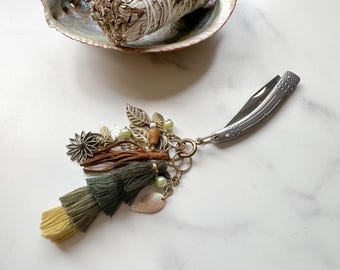 May include: A silver-toned folding pocket knife attached to a decorative keychain featuring layered tassels in olive green and yellow, brown leather strips, metal leaf charms, a flower charm, and small light green beads on a brass-colored ring.