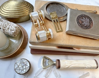 May include: A collection of vintage items including a brass shell-shaped box, a pair of gold-toned opera glasses with mother-of-pearl accents, a brass bookmark, a silver cigarette case with a nature-themed design, and a bottle opener with a carved bone handle.
