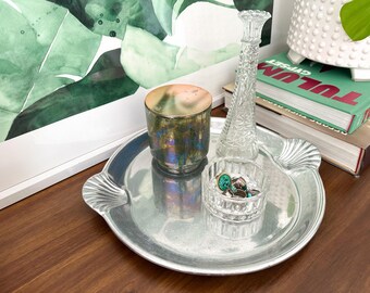 May include: A silver-colored metal tray with shell-shaped handles holds a small glass bowl with jewelry, a decorative iridescent glass, and a clear glass vase. A framed watercolor painting of green leaves is in the background.