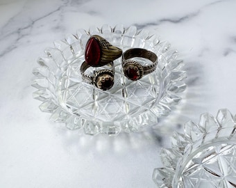 May include: Clear glass trinket dish with a starburst pattern, holding three rings. The rings have silver bands and red gemstone settings. The dish has a scalloped edge and sits on a white marble surface.