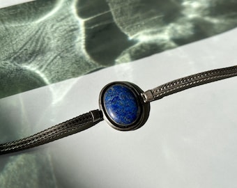 May include: A bracelet featuring a central oval-shaped blue lapis lazuli stone set in a silver-toned metal bezel. The stone is deep blue with subtle natural flecks. The band consists of a woven silver-toned chain that connects to the central setting.