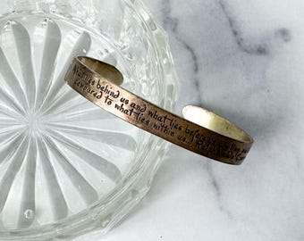 May include: A silver and copper-toned cuff bracelet with an engraved quote. The text reads "What lies behind us and what lies before us compared to what lies within us..." The bracelet is resting on a clear, patterned glass dish.