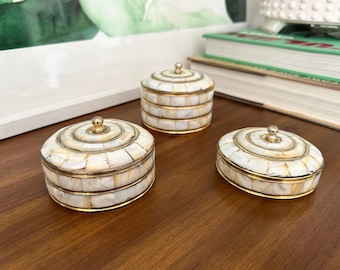 May include: Three decorative trinket boxes with mother-of-pearl inlay and gold-colored trim. The boxes are different shapes and sizes, with a round, oval, and cylindrical design. Each box has a small, gold-colored knob on the lid.