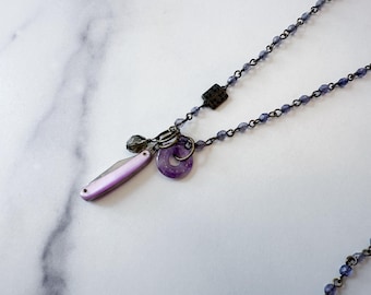 May include: A necklace featuring a chain of small, faceted purple beads and a dark metal link. The pendant consists of a small, closed folding knife with a light purple handle, a dark gray bead, and a circular, translucent purple stone ring.