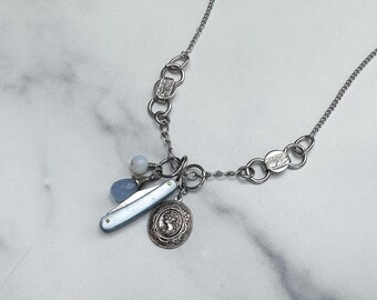 May include: A silver-toned chain necklace featuring a series of circular links and a cluster of charms. The charms include a small light blue bead, a teardrop-shaped blue stone, a miniature folding pocket knife, and a round silver medallion.