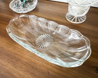 May include: An elongated, clear glass dish with a scalloped edge and a central sunburst pattern. The oval-shaped vessel features smooth, curved sides and a transparent finish, showcasing a detailed pressed glass design on the interior base.