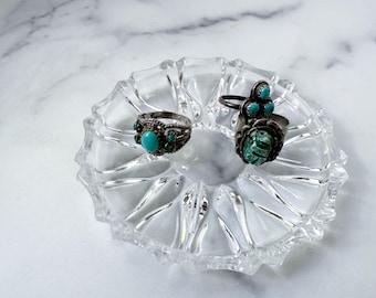 May include: A clear, glass trinket dish with a scalloped edge holds two silver rings. One ring features a large turquoise stone, while the other has multiple smaller turquoise stones. The rings are displayed on a white marble surface.