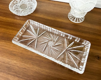 May include: A rectangular clear glass tray featuring a pressed starburst pattern across the surface and a notched rim. The tray is accompanied by a small circular glass dish and a stemmed glass bowl, all displaying intricate geometric designs.