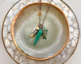 May include: A copper-toned necklace with three charms: a small folding knife, a green triangular pendant, and a green fish pendant. The necklace is displayed on a decorative plate with a gold rim and a mother-of-pearl outer edge.
