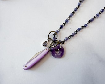 May include: A necklace featuring a chain of small purple faceted beads and a bronze-toned metal link. The pendant consists of a small purple teardrop charm, a circular purple stone ring, and a folding pocket knife with a smooth light purple handle.