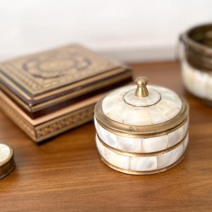 May include: A collection of decorative boxes and containers. The items include a small, round box with a lid, a round box with a lid, a rectangular box, and a round open container. The boxes have gold-colored metal trim and are made of wood and mother-of-pearl.