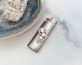 May include: A silver-toned metal lighter featuring an embossed decorative border and a cluster of iridescent, multi-faceted rhinestones arranged in a curved pattern on the front surface, with a red ignition button and a metallic top mechanism.