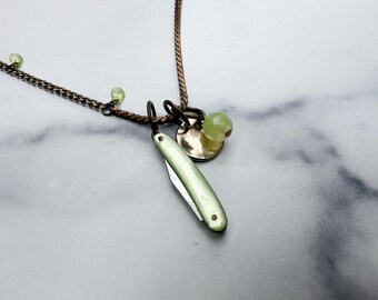 May include: A copper-toned chain necklace featuring a small, pale green pocket knife charm, a round metallic disc, and several small, faceted light green beads attached along the length of the chain. The necklace has a dark, oxidized finish.