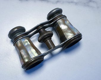 May include: Antique opera glasses with a mother-of-pearl finish. The binoculars have a dark metal frame and a central focus knob. The lenses are surrounded by a silver-colored metal. The mother-of-pearl has a pearlescent sheen.