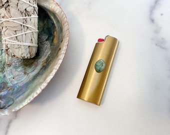 May include: A gold-toned rectangular lighter featuring a small, oval-shaped green speckled stone embedded on its front surface. The lighter has a red ignition button at the top and is positioned next to a white sage bundle in an iridescent abalone shell.