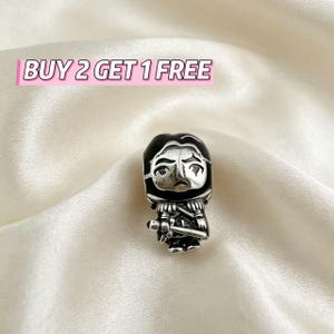 Può includere: Un ciondolo in metallo color argento raffigurante un personaggio stilizzato con dettagli in smalto nero, che indossa un indumento con cappuccio e impugna una spada. Il testo BUY 2 GET 1 FREE è visualizzato in rosa e bianco sopra il ciondolo.