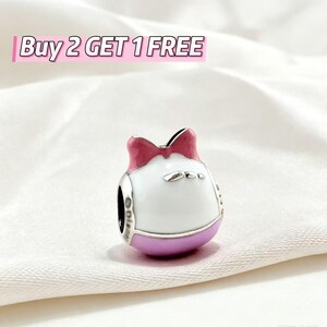 May include: A silver-toned charm bead featuring a white enamel center, a pink bow at the top, and a purple base. The charm has a central hole for threading onto a bracelet. The text Buy 2 GET 1 FREE is displayed in the upper left corner.