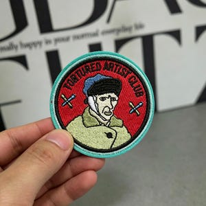 Puede incluir: Parche bordado con un retrato de Vincent van Gogh y el texto "TORTURED ARTIST CLUB". El parche tiene un fondo rojo, un borde turquesa e incluye dos pinceles cruzados. El diseño es circular.