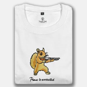 Puede incluir: Camiseta blanca con un bordado de una ardilla de dibujos animados sosteniendo un rifle. La ardilla es marrón y beige, con el texto "Peace is overrated" debajo. La camiseta tiene cuello redondo y la marca "Trand Set" es visible.