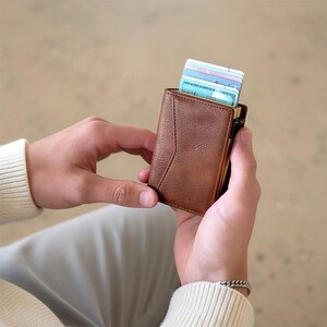 May include: A brown leather wallet with a geometric design is held open, revealing several credit cards. The wallet is a warm, medium brown colour and appears to be made of textured leather. The cards are visible at the top of the wallet.